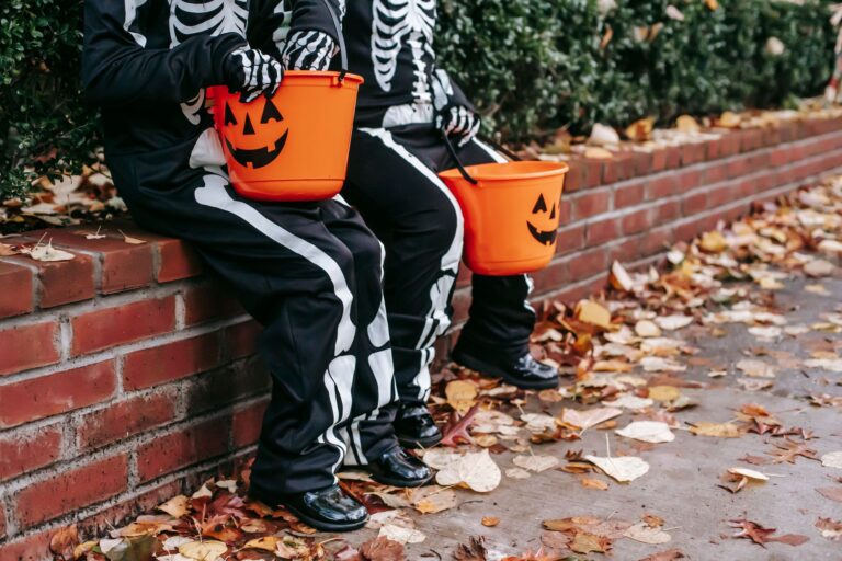 Kinder in Skelettkostümen sitzen mit Halloween-Eimern auf roter Backsteinmauer